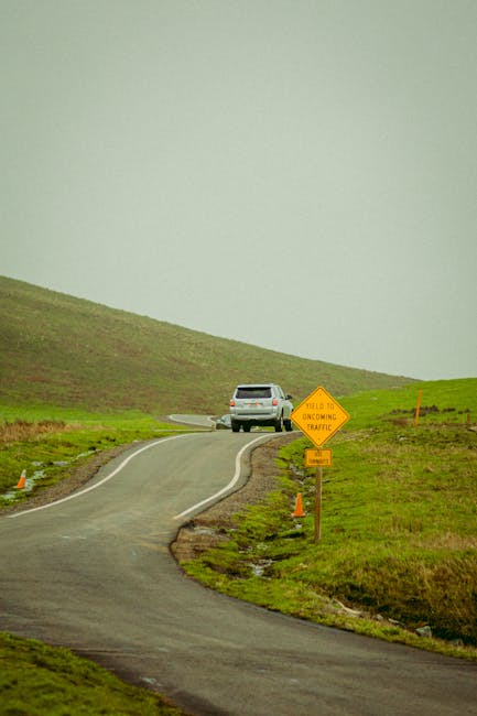 A white SUV is shown parked at the edge of a narrow, winding country lane on a grassy hillside during daytime with overcast weather. The road appears to be in a rural area with no visible buildings or houses nearby. Adjacent to the vehicle, there are two orange traffic cones placed on the pavement, and a yellow road sign indicating 'YIELD TO ONCOMING TRAFFIC' is situated beside the roadside. The scene exemplifies a quiet, scenic route that could be relevant for home relocation or moving logistics in a rural area, with the location and road conditions potentially affecting vehicle and furniture transport. The image captures the quiet environment suitable for crew-based house removals, highlighting the importance of parking and access considerations during a home move. The overall setting reflects the need for careful planning in rural moving scenarios, with the company Man with Van Crews Hill often assisting clients with such relocations.