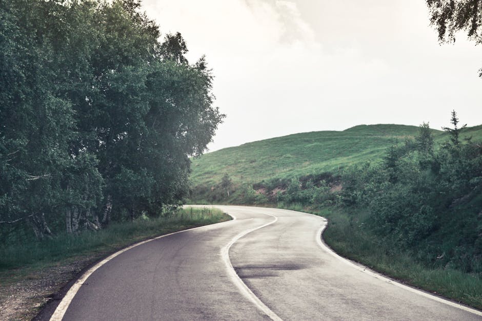 A white SUV is shown parked at the edge of a narrow, winding country lane on a grassy hillside during daytime with overcast weather. The road appears to be in a rural area with no visible buildings or houses nearby. Adjacent to the vehicle, there are two orange traffic cones placed on the pavement, and a yellow road sign indicating 'YIELD TO ONCOMING TRAFFIC' is situated beside the roadside. The scene exemplifies a quiet, scenic route that could be relevant for home relocation or moving logistics in a rural area, with the location and road conditions potentially affecting vehicle and furniture transport. The image captures the quiet environment suitable for crew-based house removals, highlighting the importance of parking and access considerations during a home move. The overall setting reflects the need for careful planning in rural moving scenarios, with the company Man with Van Crews Hill often assisting clients with such relocations.