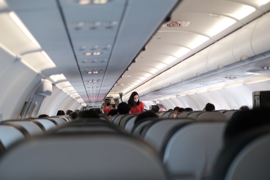 Inside the cabin of an airplane during a home relocation, showing rows of beige and gray aircraft seats with overhead storage compartments above. Several passengers are seated, with a flight attendant dressed in a red uniform and wearing a face mask standing in the aisle near the middle of the image, possibly assisting or preparing for service. The cabin lighting is soft, and the aisle extends towards the front of the aircraft, indicating a boarding or in-flight scene. This environment reflects the logistics and transport aspect of packing and moving, similar to furniture transport and loading processes that are part of house removals conducted by Man with Van Crews Hill, especially relevant during complex moving projects.