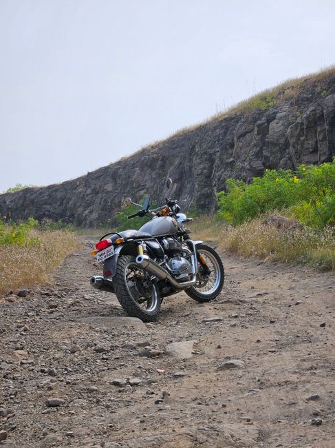 A silver and black motorcycle parked on a rough, unpaved dirt pathway with uneven rocks and loose gravel, surrounded by low green bushes and yellow wildflowers on both sides. In the background, a high