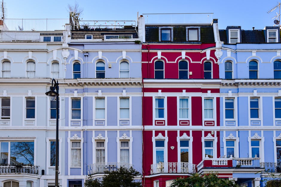 A row of three Victorian-style terraced homes with ornate architectural details and large sash windows, painted in pastel shades of blue and vibrant red; the middle house features a small balcony with white balustrades, and a black street lamp is positioned outside on the pavement. The rooftops include skylights and satellite dishes, with a clear blue sky in the background. This exterior scene depicts the urban environment where a home relocation process may take place, managed by Man with Van Crews Hill, involving careful loading and transport of furniture and boxes through the front doors or via the street for house removals or furniture transport services.