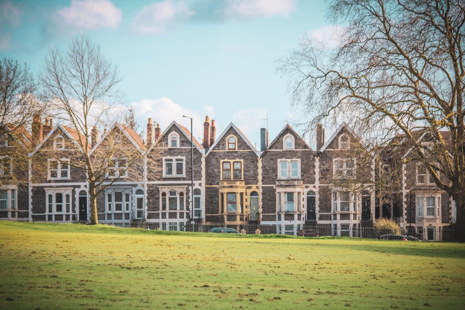 Photograph of a row of Victorian terraced homes constructed with grey brick in Crews Hill, with pitched roofs and multiple chimneys, situated behind a well-maintained grassy area and leafless trees. The homes feature large sash windows, some with white frames, and front doors of varying dark colors. The scene is captured during daylight with a clear blue sky and a few clouds. In the foreground, there are no moving objects, but the overall setting suggests an environment suitable for home relocation or furniture transport services, aligning with the scope of house removals offered by Man with Van Crews Hill. The image conveys a residential area typical for moving or packing and moving logistics, with a peaceful neighborhood context.