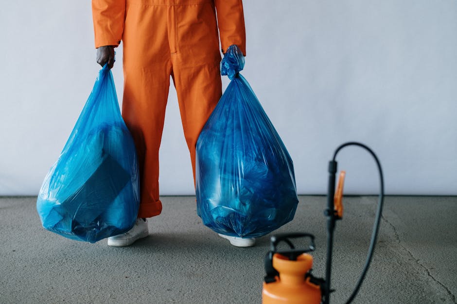 A person dressed in an orange high-visibility work uniform, including a safety helmet and gloves, is seen from above collecting and sweeping up litter from a curbside area next to a concrete wall. The