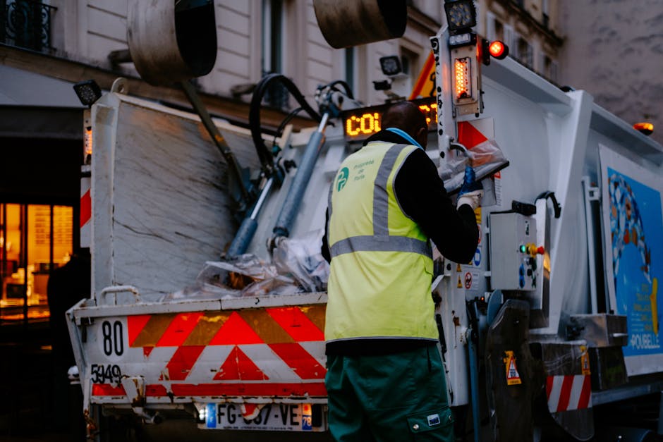 A worker wearing a high-visibility yellow vest and dark clothing is operating the control panel of a large garbage or recycling collection vehicle, which is parked outdoors near a building with illuminated windows. The vehicle has red and white diagonal safety markings on its rear, and the worker is seen from behind, adjusting controls or inspecting the machine. Inside the truck's open container, visible materials include cardboard boxes, plastic wrapping, and piled waste, indicating a collection or disposal process. The scene is set during the evening or night, with artificial lighting illuminating the area. This image captures a residential or urban setting where waste collection is in progress, aligning with home relocation logistics and waste management aspects relevant to moving services provided by Man with Van Crews Hill.