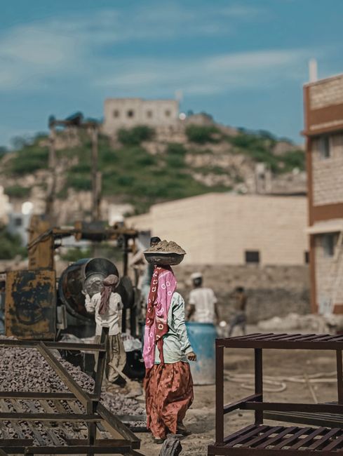 A woman wearing a wide-brimmed hat, a pink scarf draped over her shoulders, a light-colored top, and a long skirt, stands near a construction site with a large yellow excavator. She is facing away from the camera, observing or working at the site. Several other workers can be seen in the background, some wearing white shirts and head coverings, engaged in manual labour around piles of gravel and construction equipment. The setting is outdoors with a partially built or unfinished brick structure on the right side of the image. In the distance, a hill with a fortress or church on top is visible under a partly cloudy sky. The scene depicts an active area of home or building relocation, illustrating the logistics and physical effort involved in property or infrastructure development, aligning with services such as those offered by Man with Van Crews Hill for removals and moving projects.