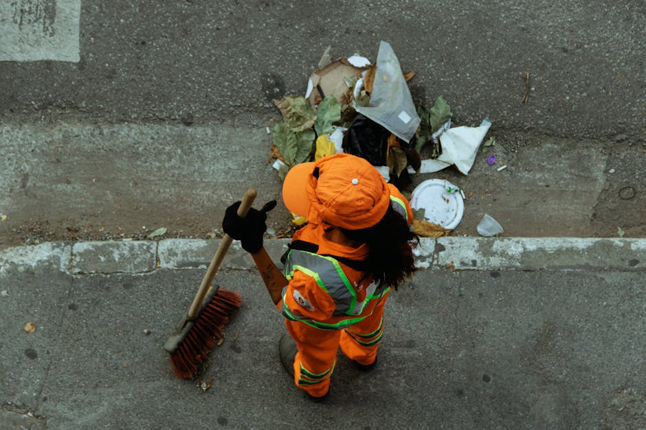 A worker wearing a high-visibility yellow vest and dark clothing is operating the control panel of a large garbage or recycling collection vehicle, which is parked outdoors near a building with illuminated windows. The vehicle has red and white diagonal safety markings on its rear, and the worker is seen from behind, adjusting controls or inspecting the machine. Inside the truck's open container, visible materials include cardboard boxes, plastic wrapping, and piled waste, indicating a collection or disposal process. The scene is set during the evening or night, with artificial lighting illuminating the area. This image captures a residential or urban setting where waste collection is in progress, aligning with home relocation logistics and waste management aspects relevant to moving services provided by Man with Van Crews Hill.