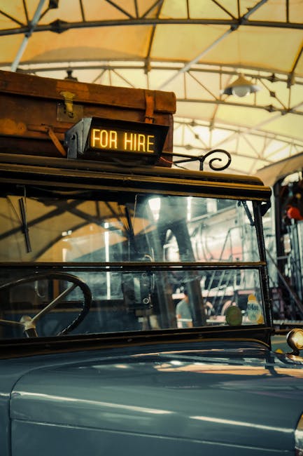 Close-up view of a vintage vehicle parked outdoors under a canopy, with a black digital sign mounted on the roof displaying 'FOR HIRE' in bright yellow-orange letters. The vehicle features a glass win