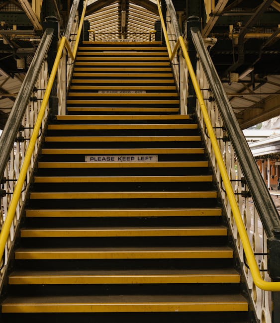 A close-up view of a metal staircase with black and yellow striped anti-slip treads, installed inside a warehouse or loading area. The staircase features yellow handrails on both sides, supporting saf