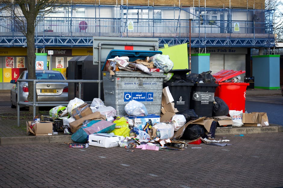 A large collection of overflowing waste and recycling bins situated on a paved sidewalk outside a commercial building in Crews Hill, Enfield. The waste includes black, red, and green bins, with some lids open and items such as cardboard boxes, plastic bags, paper, and packaging materials spilling onto the ground. The surrounding area features a metal railing, parked silver car, and a store facade with signs and promotional posters. In the background, there is a building under construction or renovation, covered with blue scaffolding and tarpaulin. The scene illustrates improper waste disposal, which can impact local waste management regulations and house removal planning. Man with Van Crews Hill offers professional removals and moving services, assisting with waste clearing and logistical coordination for home relocations, ensuring compliance with local waste rules.