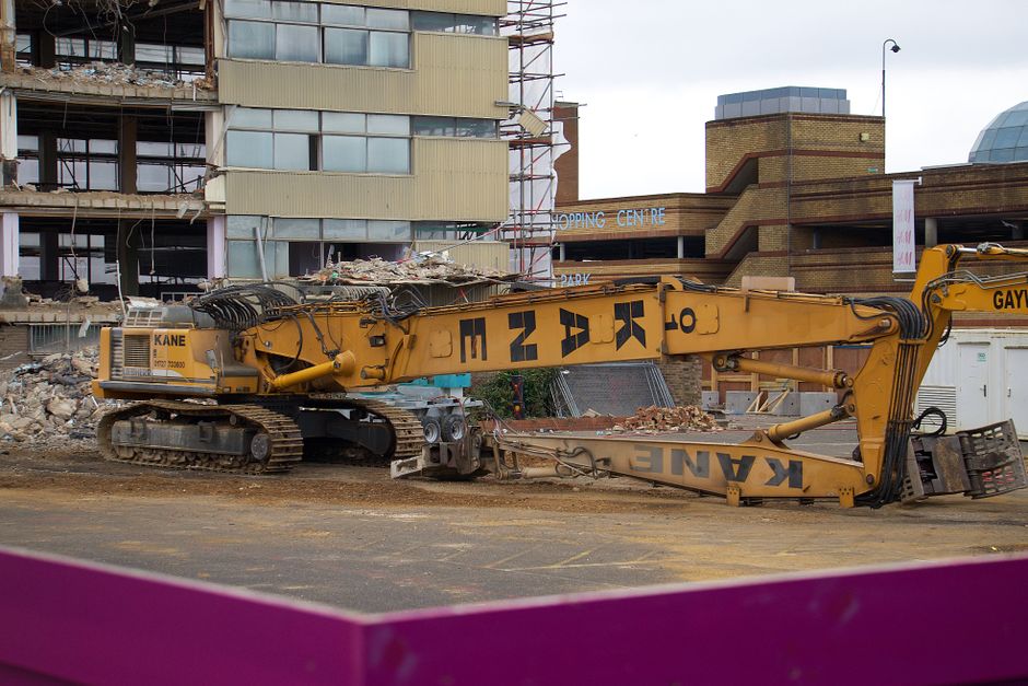 A large yellow demolition excavator with tracked wheels and extended hydraulic arm is situated on a construction site in Crews Hill under Enfield. The excavator's arm is resting on the ground, with the bucket attachment tilted downward, holding debris from a partially demolished building. The building in the background is under renovation or destruction, with exposed concrete, metal framework, and debris scattered around. Behind the site, there is a multi-storey structure with glass windows and a brick facade, and a sign indicating a shopping centre. The scene is set during daytime with overcast sky, and the site appears to be in the process of home relocation or building clearance, for which Man with Van Crews Hill offers removals and moving services.