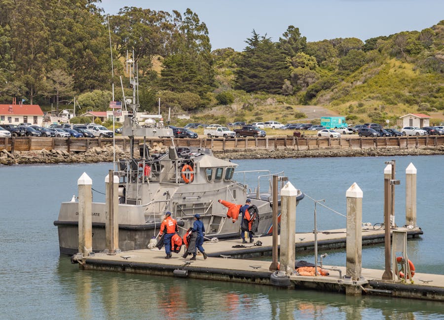 A group of four crew members wearing waterproof jackets and backpacks are standing on a floating dock, preparing to load a grey patrol boat onto the vessel. The boat is moored to wooden pylons on a calm body of water, with some crew members actively lifting or guiding a large orange bag or equipment onto the boat. In the background, there is a shoreline with parked cars, trees, and small buildings, indicating a coastal area. The scene is set during daytime with clear weather, and the boat is equipped with antennas, radar, and communication devices. This image captures the process of equipment transfer or maintenance related to marine operations, with visible safety gear and procedural activity that could be part of a home relocation or boat transport service provided by Man with Van Crews Hill.