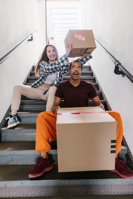 Two young individuals, a woman and a man, sit on a staircase inside a building during a home relocation process. The woman, wearing a checkered shirt and beige trousers, is smiling and holding a medium-sized cardboard box above her head. The man, dressed in a maroon t-shirt and bright orange trousers, is seated on the stairs holding a larger cardboard box on his lap. The staircase is carpeted with dark grey material and has metal handrails on both sides. The background features a white-painted wall and a door opening to an upper level or outside area, with natural light illuminating the scene. The image captures the act of packing and moving, with the boxes wrapped in clear plastic and packing tape, reflecting the packing and moving services provided by Man with Van Crews Hill, supporting efficient furniture transport and home relocation.